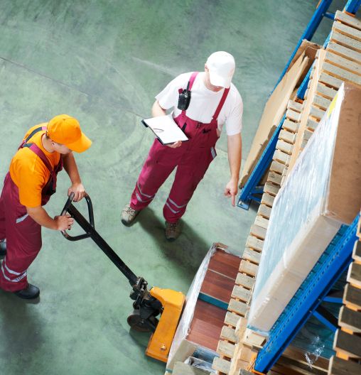 Zwei Arbeiter in dunkelroten Overalls räumen ein Lager ein. Einer der beiden betätigt einen Gabelstapler.